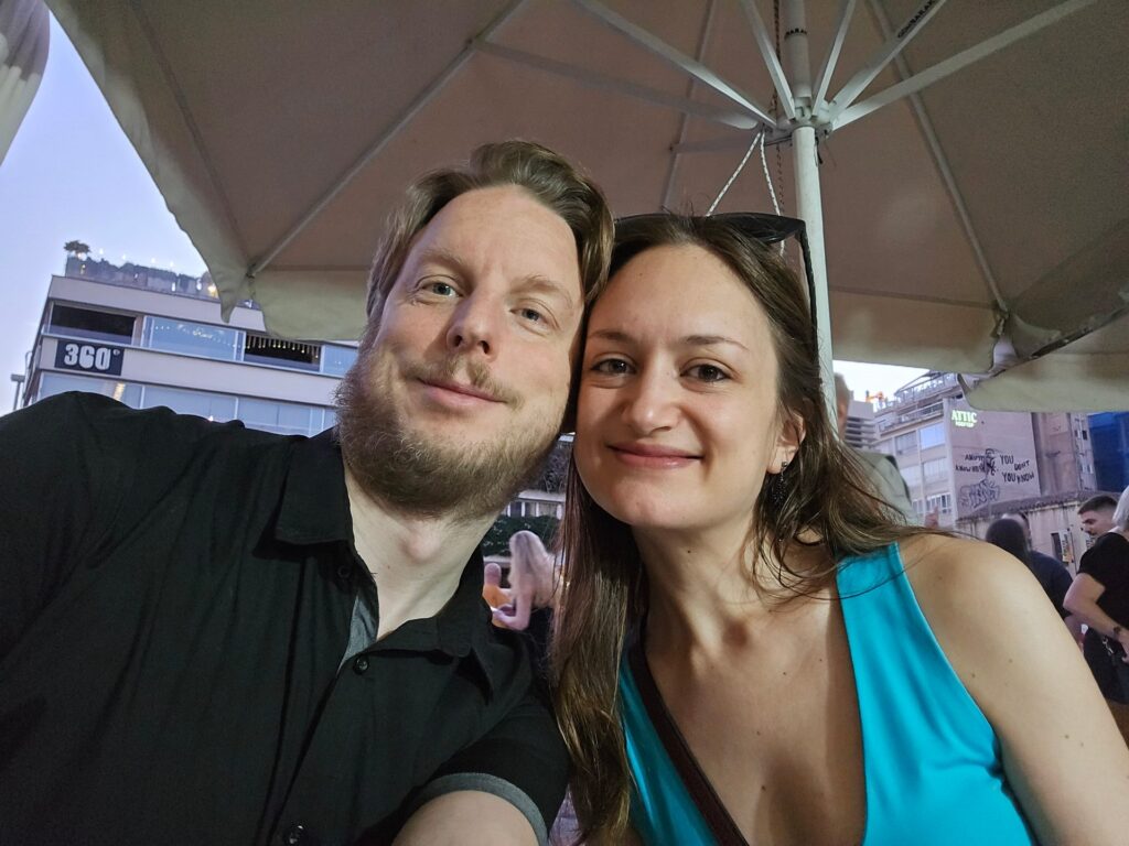 Florian und Eliana lächelnd unter einem Sonnenschirm in Athen